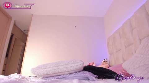 Video of a woman in a white nightgown lying on a bed, surrounded by various pillows, in a dimly lit bedroom with beige walls and a textured headboard.