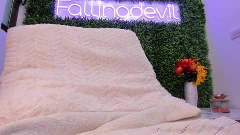 A video of a cozy room with a plush, cream-colored blanket draped over a chair. Behind, a vibrant green wall with \"Follinrobevil\" neon sign. A vase with yellow and red flowers sits on the right.