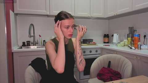 Video of a young woman with pale skin and dark hair in a messy bun, wearing a black sleeveless top, sitting at a kitchen table. She's holding her glasses, appearing surprised. The kitchen has white cabinets and a stainless steel sink.