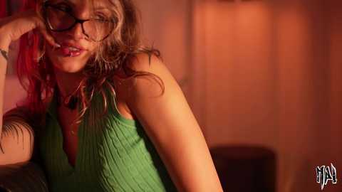 Video of a blonde woman with red highlights in her hair, wearing black-rimmed glasses, a green ribbed tank top, and a necklace. She leans against a warm-colored wall, with a blurred background.