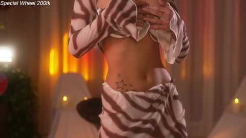 A video of a woman with light skin and toned abs, wearing a white striped crop top and matching sarong, with a star tattoo on her hip, standing in a warmly lit room.
