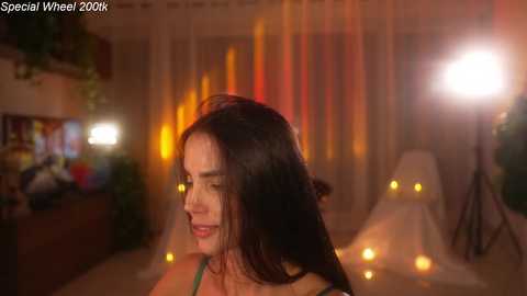 Video of a young woman with long dark hair, fair skin, and a small frame, smiling gently. She's in a dimly lit, warmly decorated room with glowing candles and red and orange lighting.