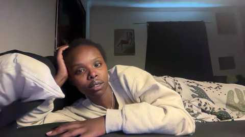 A video of a young Black woman with short hair lying on a bed, wearing a light-colored robe, looking sad. The room has dark curtains and a painting on the wall.