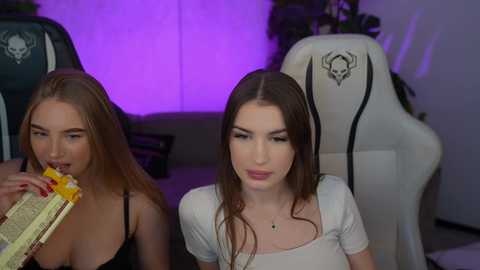 Video of two young women with long hair, one eating cake, seated in gaming chairs, in a dimly lit room with purple lighting.