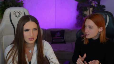 Media: Video of two women in a modern living room: brunette in a white shirt, redhead in black top, both seated on gaming chairs.