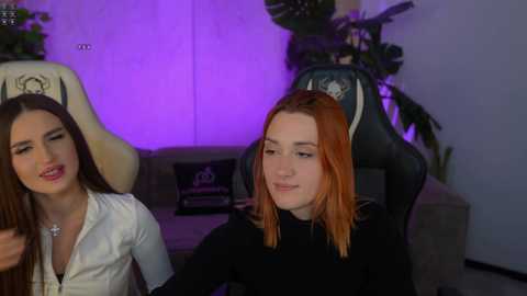 Media: Video of two young women with straight hair, one brunette, one red, sitting on a gray couch. Purple lighting, plants, and a gaming chair in the background.