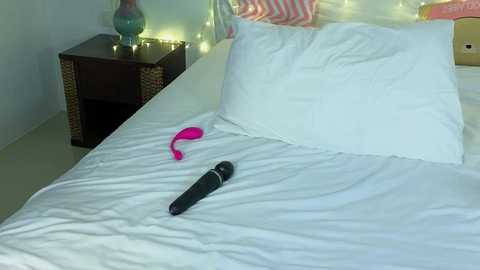 Video of a neatly made bed with white sheets, a pink vibrator, and a black vibrator on the mattress, against a backdrop of a dark wooden nightstand with a decorative lamp and colorful pillows.