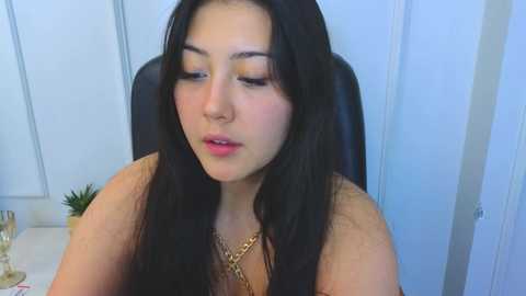 Video of a young woman with long black hair and fair skin, wearing a gold necklace, sitting in a black chair against a white background.
