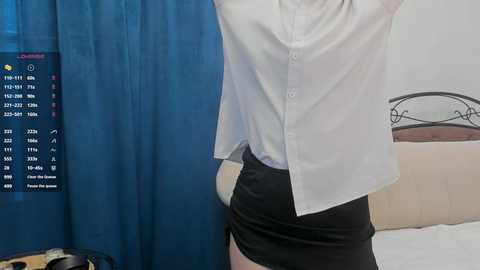 Video of a person in a white button-up shirt, black skirt, and blue curtains in a bedroom. A TV screen displays a flight schedule.