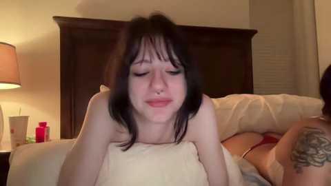 Video of a young woman with straight black hair and pale skin, lying on a bed with a beige blanket, wearing a red bra, smiling. Background includes a dark wooden headboard and a lamp on a nightstand.