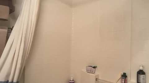 Video of a small, clean bathroom with a white-tiled shower area, featuring a white curtain, a soap dish, and several bottles of toiletries.