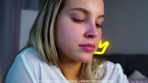 Video of a young blonde woman with fair skin, wearing a white shirt, looking contemplative. The background is blurred, featuring a gray wall and a faint yellow arrow.