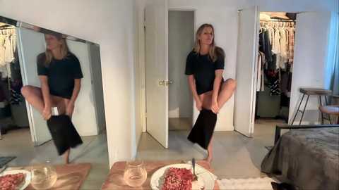 A video shows a blonde woman in a black outfit, standing in a mirror, lifting one leg, with a bed and open closet in the background.