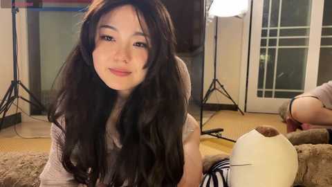 Video of a young East Asian woman with long, wavy black hair, smiling in a cozy, dimly lit living room. She wears a light-colored sweater. Background includes a lamp, plush toys, and a glass door.