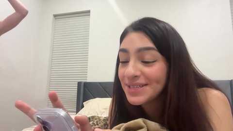 Video of a young Latina woman with long, dark hair, wearing a black top, smiling while holding a silver flip phone in a simple, beige bedroom.