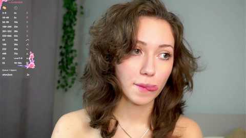 Media: Video of a young woman with wavy brown hair, light skin, and a slender build, wearing a simple necklace. Background features a computer screen displaying a calendar and green vines.