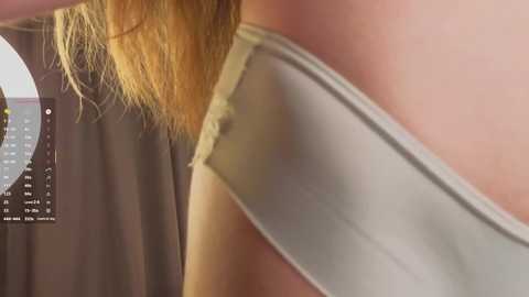 A close-up video of a person's shoulder and arm, revealing a sleeveless gray top with a slight tear, and long, blonde hair. The background features a circular digital clock displaying the time.