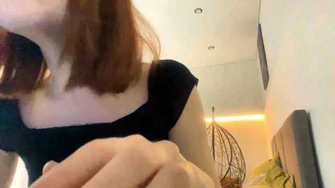 A video shows a woman with shoulder-length auburn hair, wearing a black top, bending over, and looking to the side. The background features a modern bedroom with a gray headboard, a decorative lamp, and a minimalist decor.