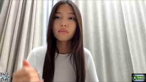 Video of a young Asian girl with long black hair, light skin, wearing a white t-shirt, standing in a room with beige curtains. She has a neutral expression.