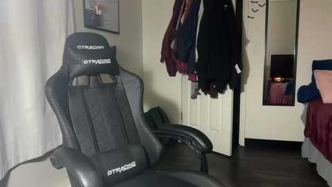 Video of a modern gaming chair with a black frame and grey cushion, positioned in a cozy bedroom with a white door, dark wooden floor, and a bed with red and blue bedding.