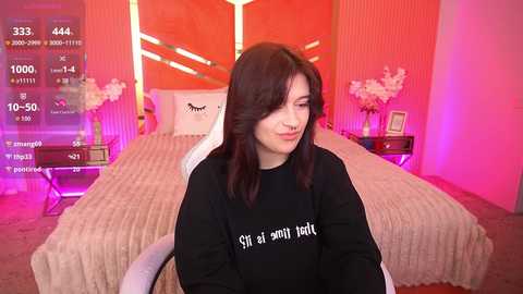 Video of a young woman with long dark hair, wearing a black sweatshirt, seated in a modern, pink-themed bedroom with a neatly made bed, pink floral arrangements, and a digital display showing a 374 temperature.