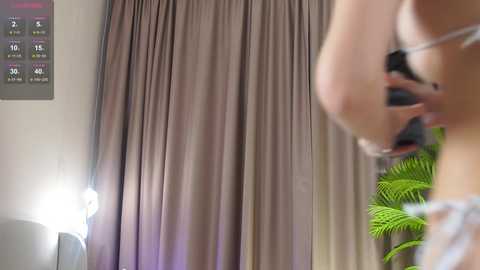A blurred video of a person with light skin and wearing a black dress, holding a camera, against a backdrop of taupe curtains and a green fern.