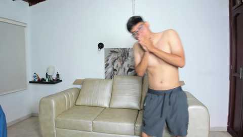 Video of a shirtless, thin Asian man with glasses, wearing gray sweatpants, standing in a minimalist living room with a beige leather sofa and a black-and-white lion painting on the wall.