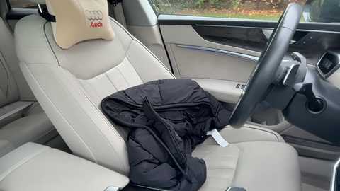 Video of a beige leather car seat with a black puffer jacket draped over it, a beige pillow with red text, and a steering wheel.