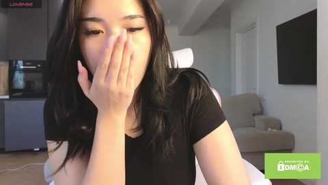Video of an Asian woman with long black hair, wearing a black shirt, covering her face with her hands, in a modern, light-colored living room with kitchen in the background.