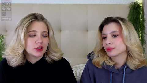 Video of two young women with blonde hair, fair skin, and light makeup. One wears a black top, the other a blue hoodie. They smile at the camera against a beige tufted headboard background.