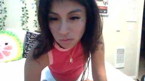 Media: Video of a young woman with medium-brown skin and long, dark hair, wearing a red tank top, indoors, surrounded by green ivy, black chalkboard, and white walls.