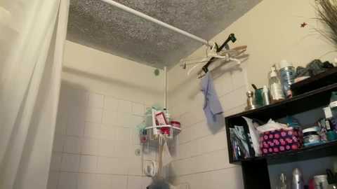 Video of a small, cluttered bathroom with white tiled walls, a white shower curtain, and various toiletries, including shampoo bottles, hanging on a rod.