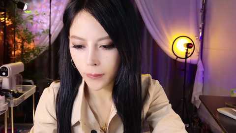 Video of a young Asian woman with long black hair, fair skin, and light makeup, seated at a sewing machine in a dimly lit room with purple and white curtains and a vintage lamp.