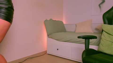 Media: A video of a small, dimly lit bedroom with a white bed, green pillows, and a black office chair. The wall is plain, and the room has a cozy, intimate feel.