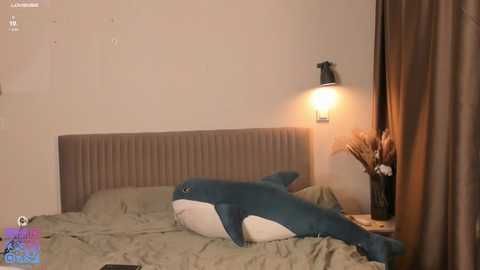 A video of a plush shark toy on a bed, with a beige headboard, beige pillows, a lamp, and a vase of dried flowers on a nightstand.