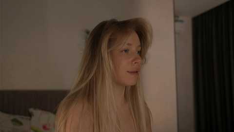 Video of a fair-skinned, blonde-haired woman with messy hair, wearing glasses, in a dimly-lit bedroom, possibly mid-30s, with a bed and dark curtains in the background.