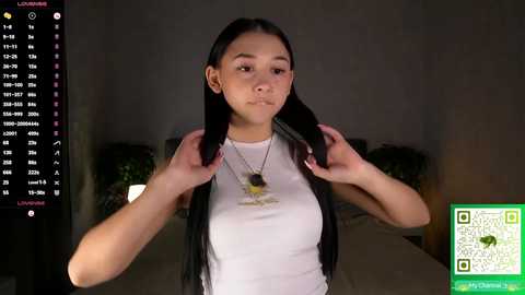 Video of a young Asian woman with long black hair, wearing a white tank top, adjusting a black headset mic, in a dimly lit room with green QR code overlay.