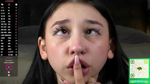 Video of a young Asian woman with long black hair, wide eyes, and a nose piercing, covering her mouth with her fingers. Background shows a calendar and QR code.