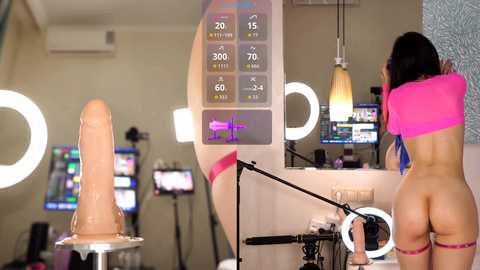 Video of a woman with long black hair, wearing a pink crop top and red thong, standing in a modern studio with a large dildo, a ring light, and monitors displaying fitness data.