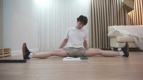 Media: Video of a young man in a white T-shirt and gray shorts, doing a wide-legged stretch on a wooden floor. Background features a beige couch, a book, and beige curtains.