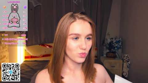 A video of a young Caucasian woman with straight, light brown hair, fair skin, and blue eyes. She is topless, sitting in a dimly lit bedroom with a bed, nightstand, and wall art. The image includes a QR code and social media icons.
