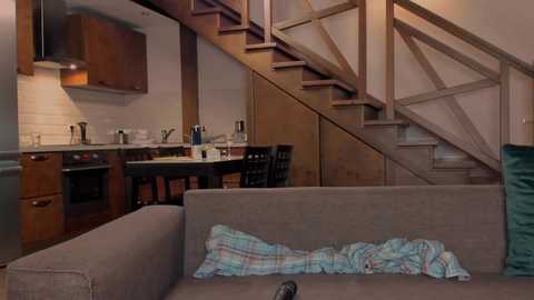 A video of a cozy, modern kitchen with wooden cabinets, stainless steel appliances, and a wooden staircase leading upstairs. A beige couch with plaid blankets and a TV remote in the foreground.