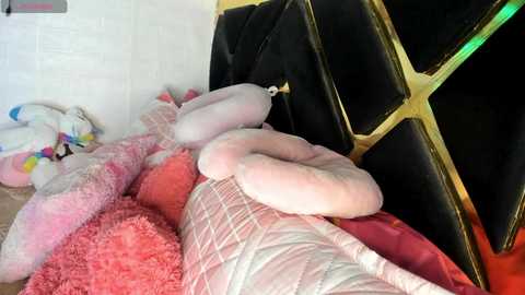 Media: Video of a cluttered, pink and white quilted bed with plush toys, including a large stuffed animal, against a black and gold geometric wall.
