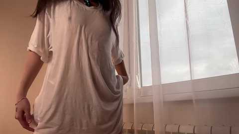 Media: Video of a woman in a loose white t-shirt, standing near a window with white blinds. She has long brown hair and is smiling.