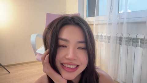 Video of an East Asian woman with long brown hair, smiling, one hand on her face, in a cozy room with a white radiator and window.