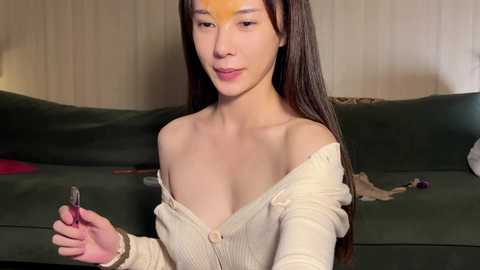 A video of a young Asian woman with long dark hair, fair skin, and medium-sized breasts, wearing a beige off-shoulder top. She holds a black lipstick, smiling in a cozy living room with a green couch and wooden walls.