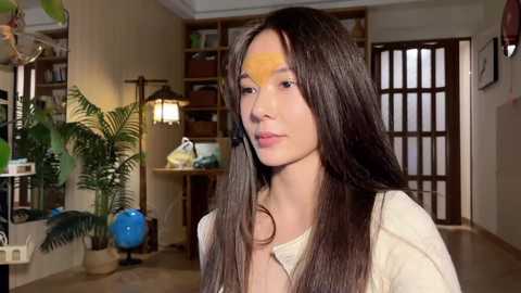 Video of a young Asian woman with long dark hair, wearing a beige top, standing in a cozy, warmly lit living room with bookshelves, a globe, and ferns.