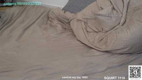 A close-up video of a bed with beige sheets, showing a wrinkled, crumpled state. The background includes a grey pillow and a partial glimpse of a wooden floor. The image is marked with a QR code and text stating \"control my toy 102.\