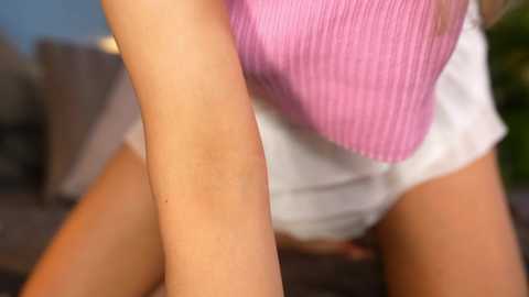 Media: A close-up video of a person's arm wearing a white sleeveless shirt and a pink, textured cardigan, seated on a dark sofa with blurred background.