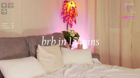 A video of a modern bedroom with a gray upholstered bed, white pillows, a vibrant, colorful lampshade, and a bouquet of white flowers on a nightstand. The background features soft, pastel-colored curtains.